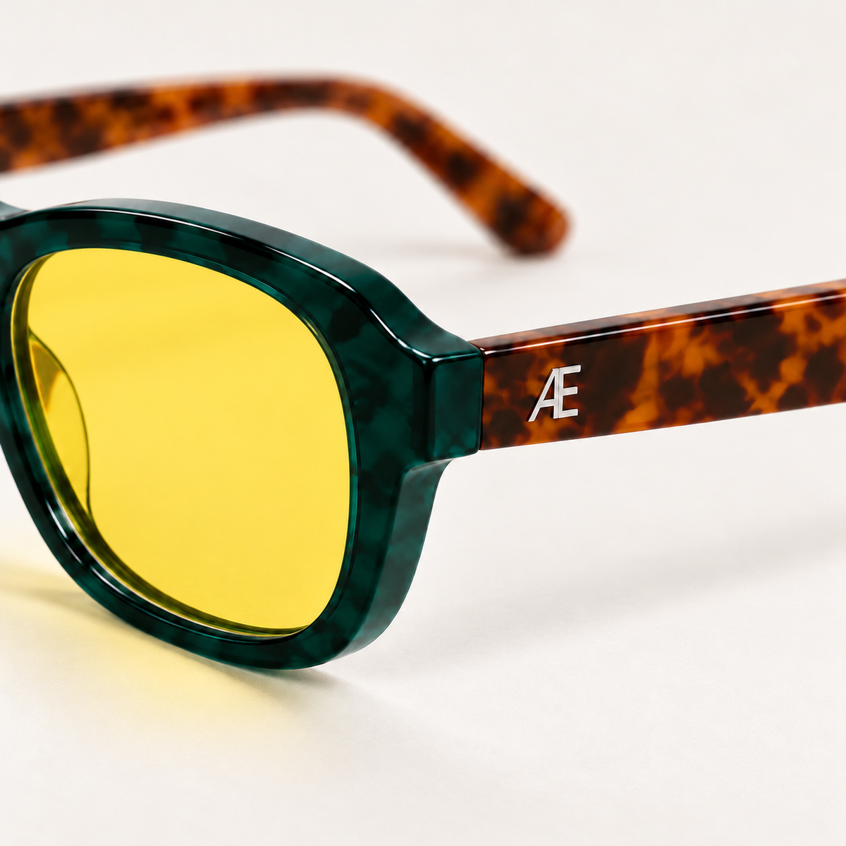 Macro close-up of ATÉN Forest/Tortoise glasses with a pronounced amber Focus Mode lens; deep forest-green rim, brown tortoiseshell outer temple and raised silver Æ monogram in focus, with the opposite arm blurred on a warm ivory background.