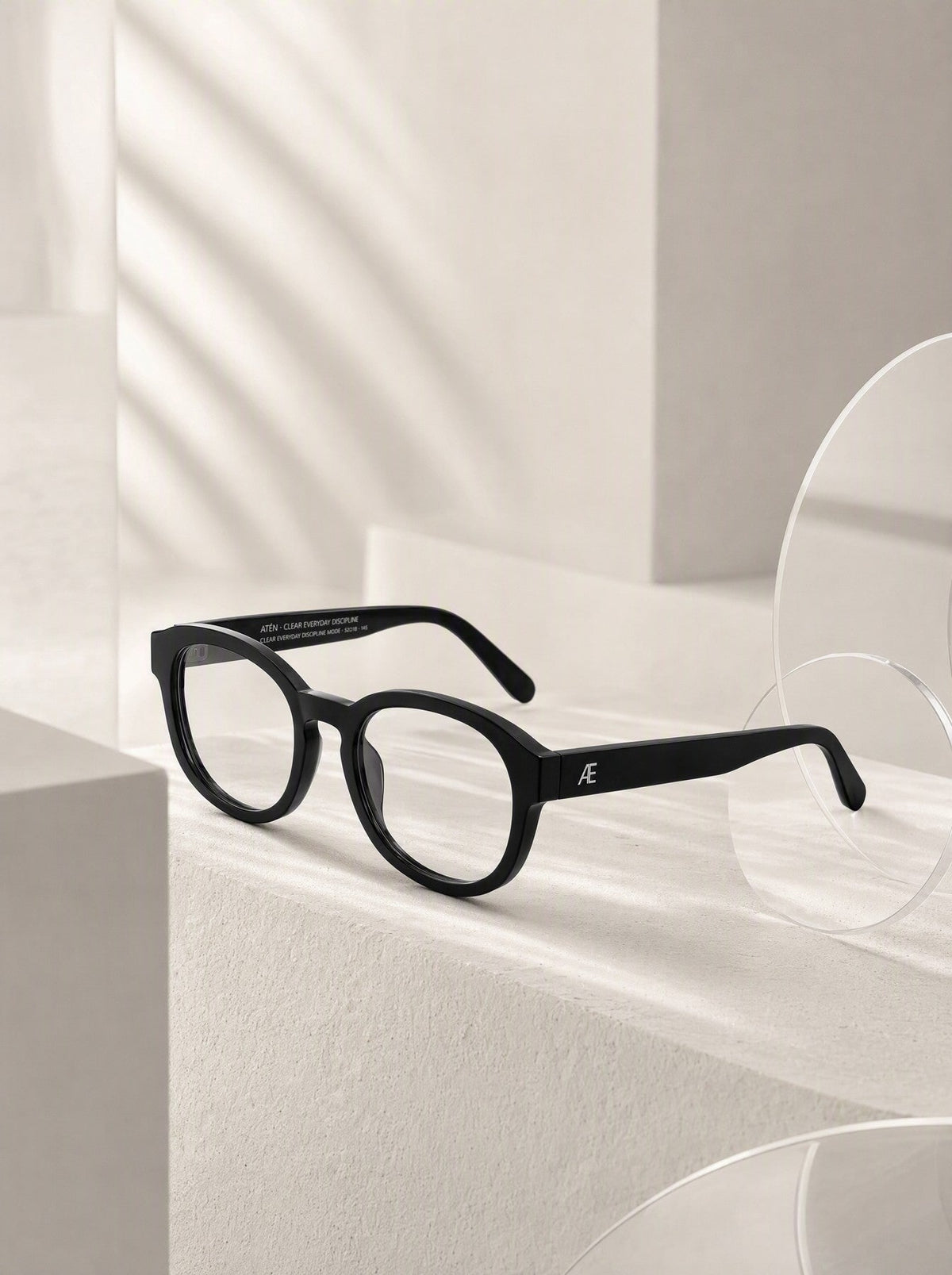 Open ATÉN Matte Black glasses with Everyday Mode lenses with a subtle warm tint in a front-right three-quarter view; silver ATÉN monogram on the near temple and small white inner-temple engraving, on a neutral studio background.
