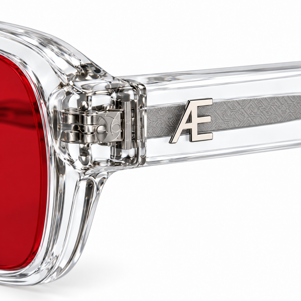 Macro of ATÉN Crystal Clear frame with deep red After Hours lens, exposed silver hinge and raised monogram on white background.