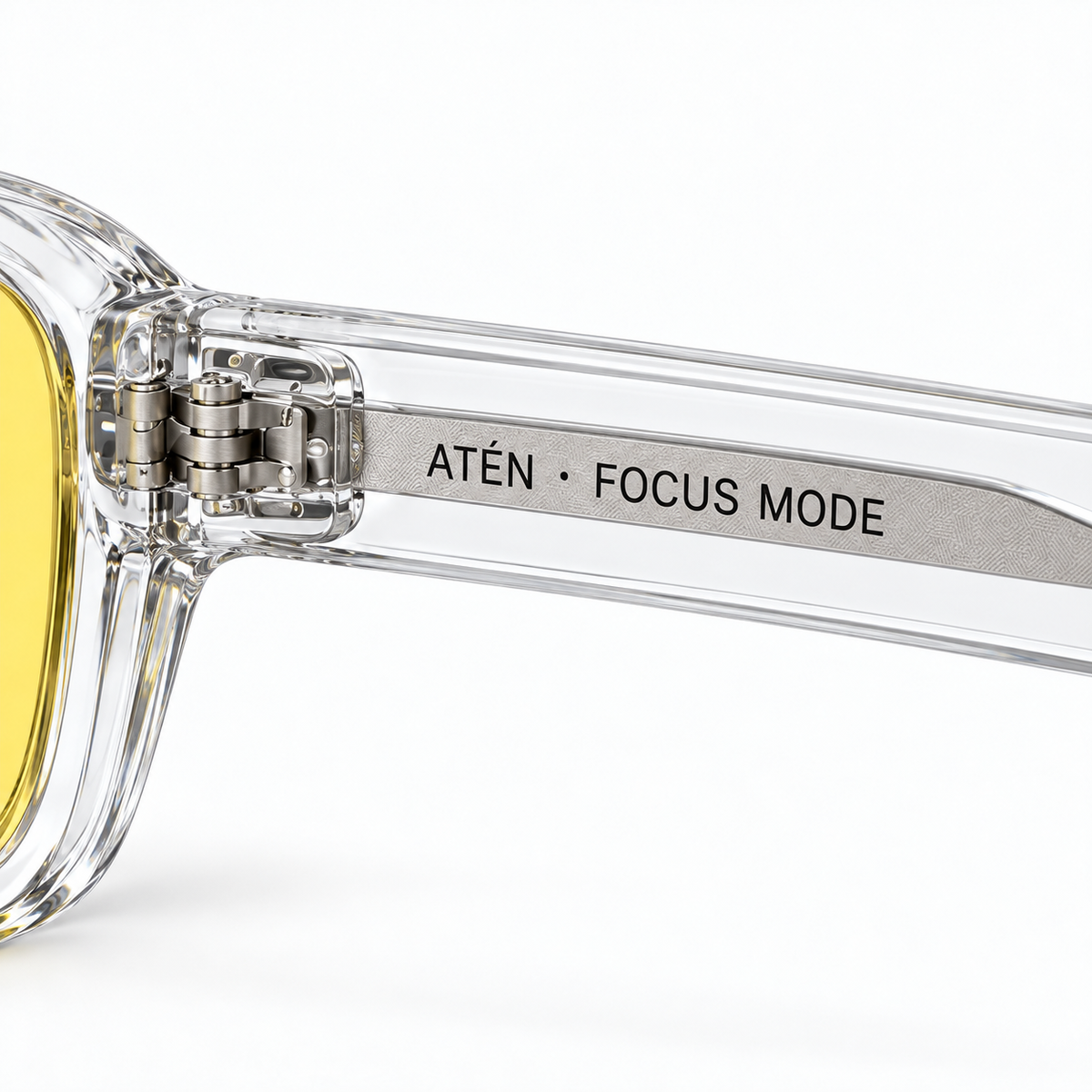 Macro of ATÉN Crystal Clear hinge, pronounced amber Focus Mode lens edge and silver core reading “ATÉN · FOCUS MODE” on white background.