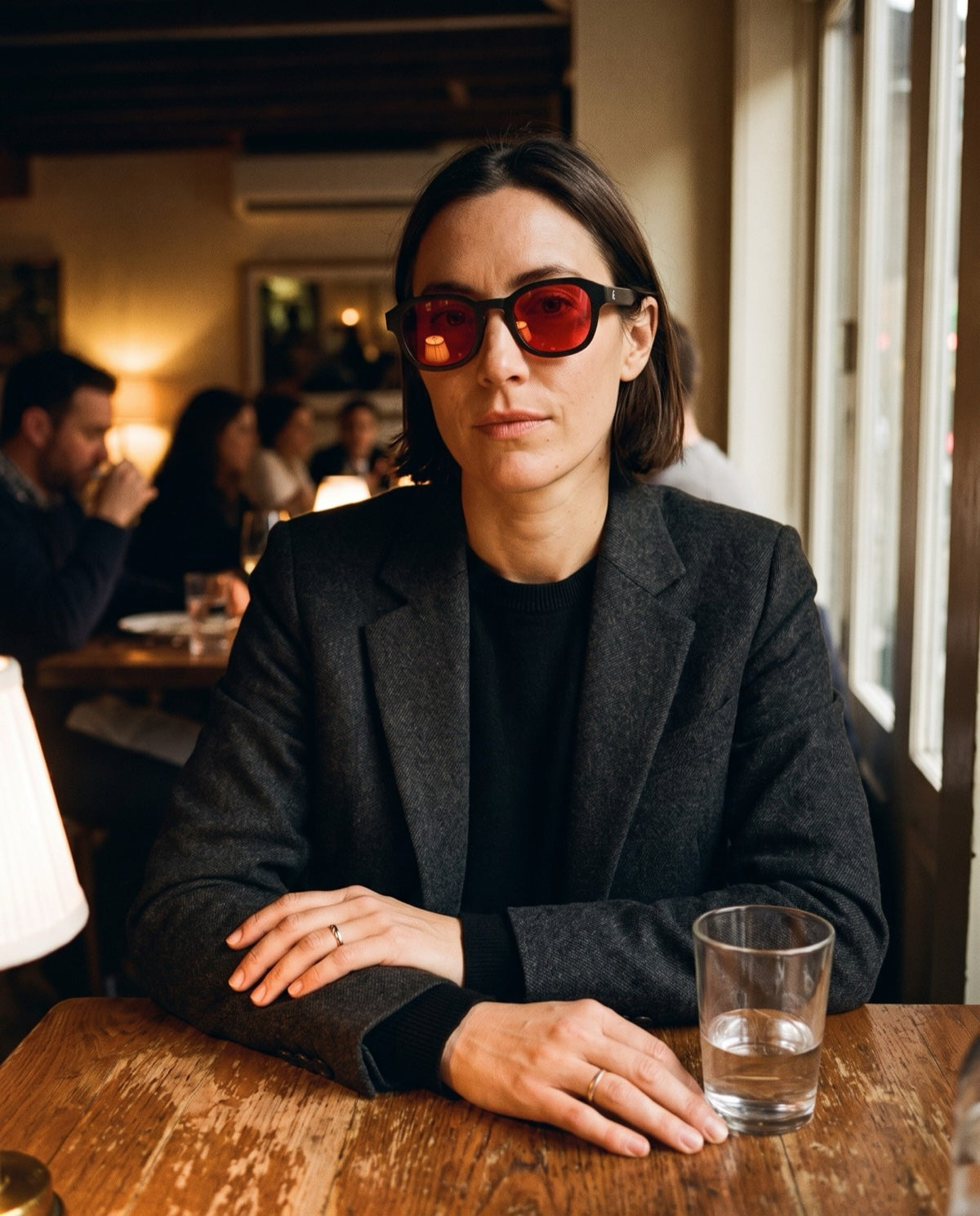 Front-facing woman with a chin-length dark-brown bob wearing black ATÉN frames with deep red After Hours lenses, seated at a worn café table in a charcoal blazer, with crossed hands, a water glass, table lamp and softly blurred patrons behind.