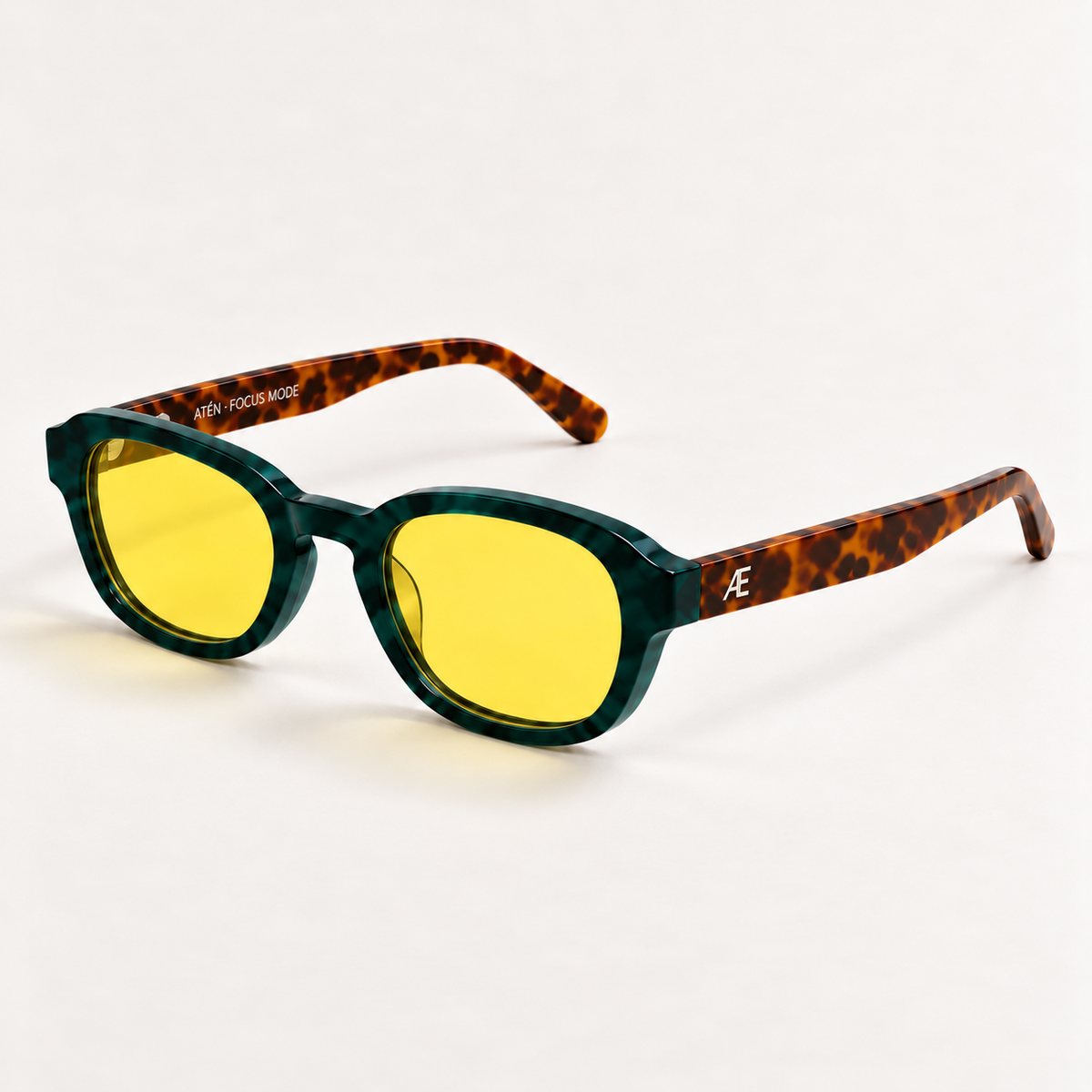 Open ATÉN Forest/Tortoise glasses with pronounced amber Focus Mode lenses in a front-right three-quarter view; deep forest-green front, brown tortoiseshell arms, silver Æ monogram and inner ATÉN · FOCUS MODE engraving on a warm ivory background.