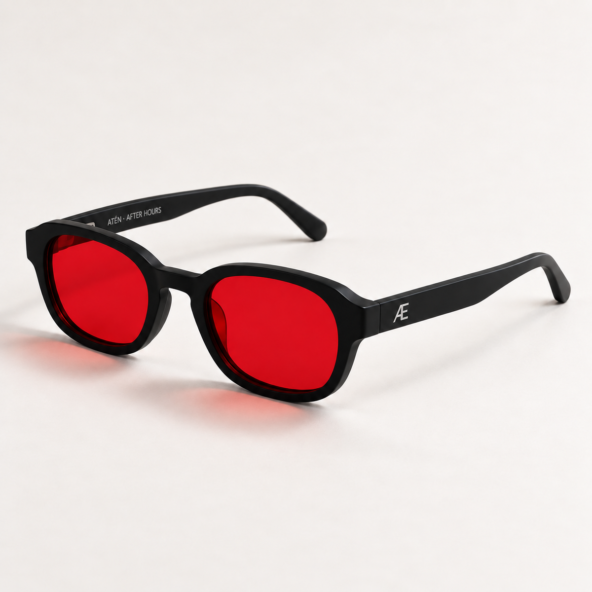 Open ATÉN Matte Black glasses with deep red After Hours lenses in a front-right three-quarter view; silver Æ monogram on the outer temple and inner ATÉN · AFTER HOURS engraving visible on a warm ivory studio surface.
