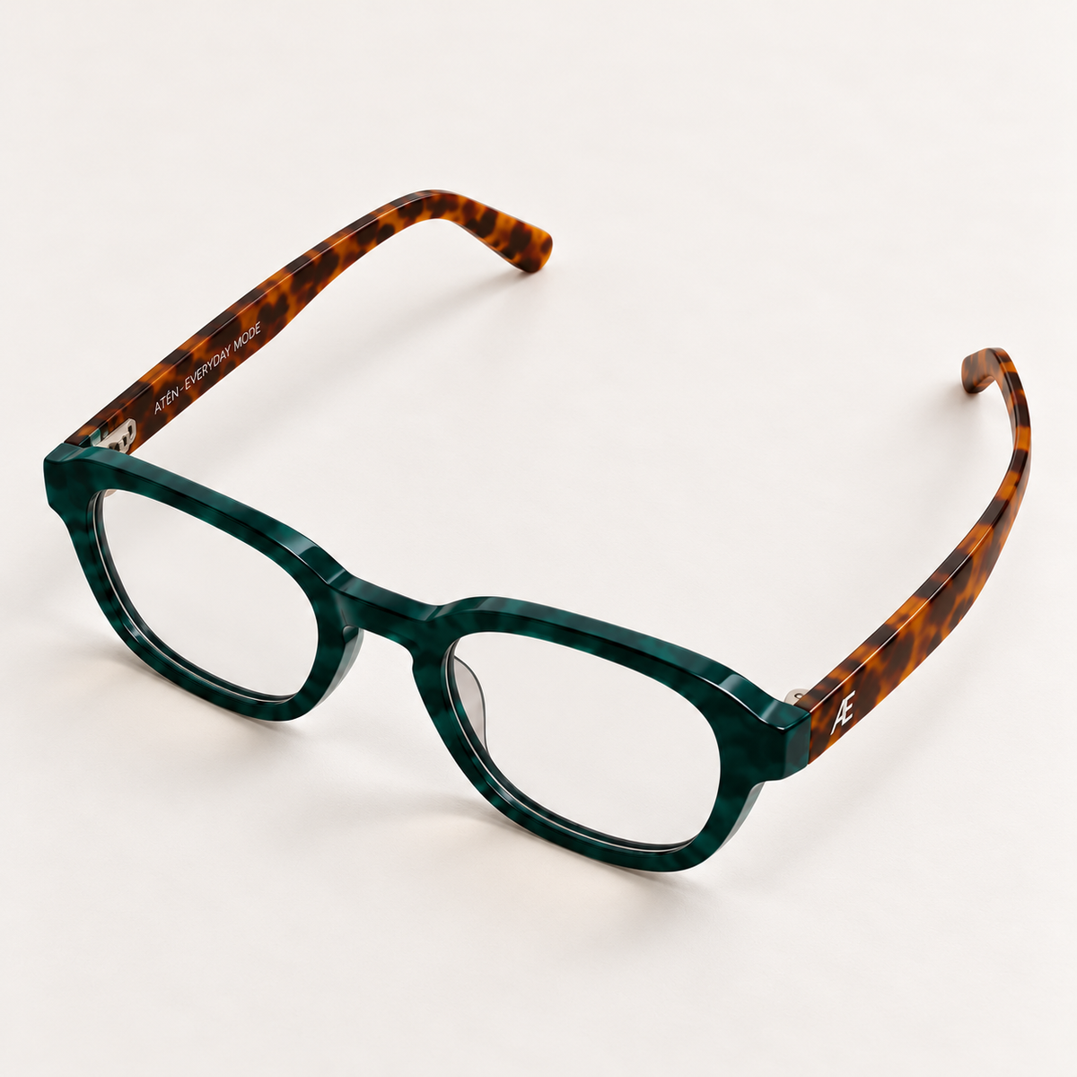 Open ATÉN Forest/Tortoise glasses with Everyday Mode lenses with a subtle warm tint, viewed from above at a front three-quarter angle; deep green front, tortoiseshell temples, silver monogram and small inner-temple engraving on ivory.