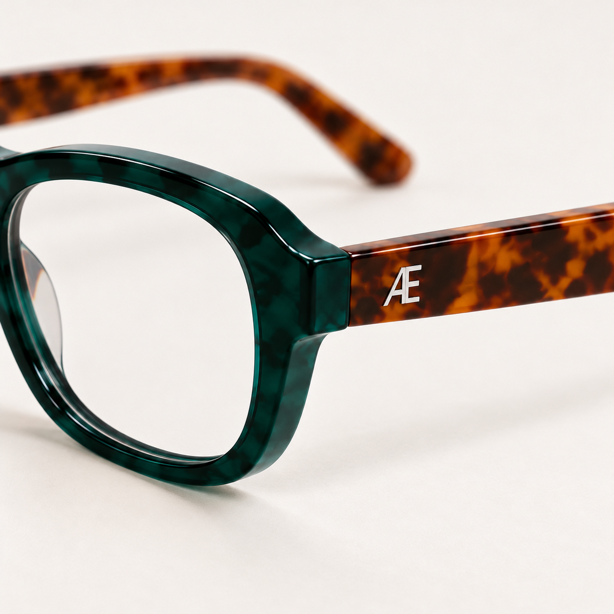 Three-quarter macro of ATÉN Forest/Tortoise glasses with an Everyday Mode lens with a subtle warm tint on ivory; green rim, brown temple and silver monogram.