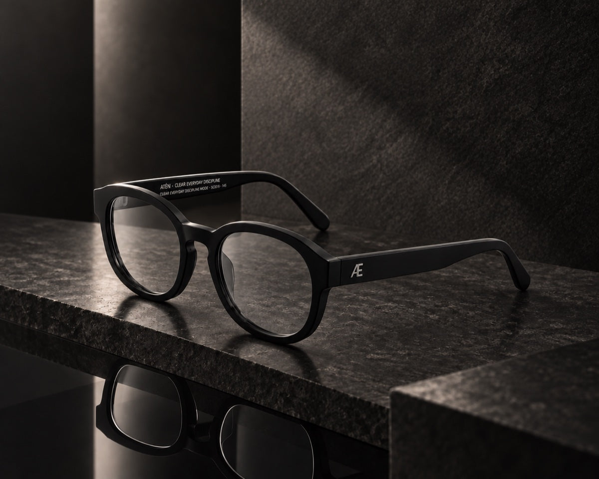Open ATÉN Matte Black glasses with Everyday Mode lenses with a subtle warm tint on a glossy charcoal stone slab, viewed from the front-right; the silver Æ temple monogram and inner-temple lettering are visible in hard side light and reflection.