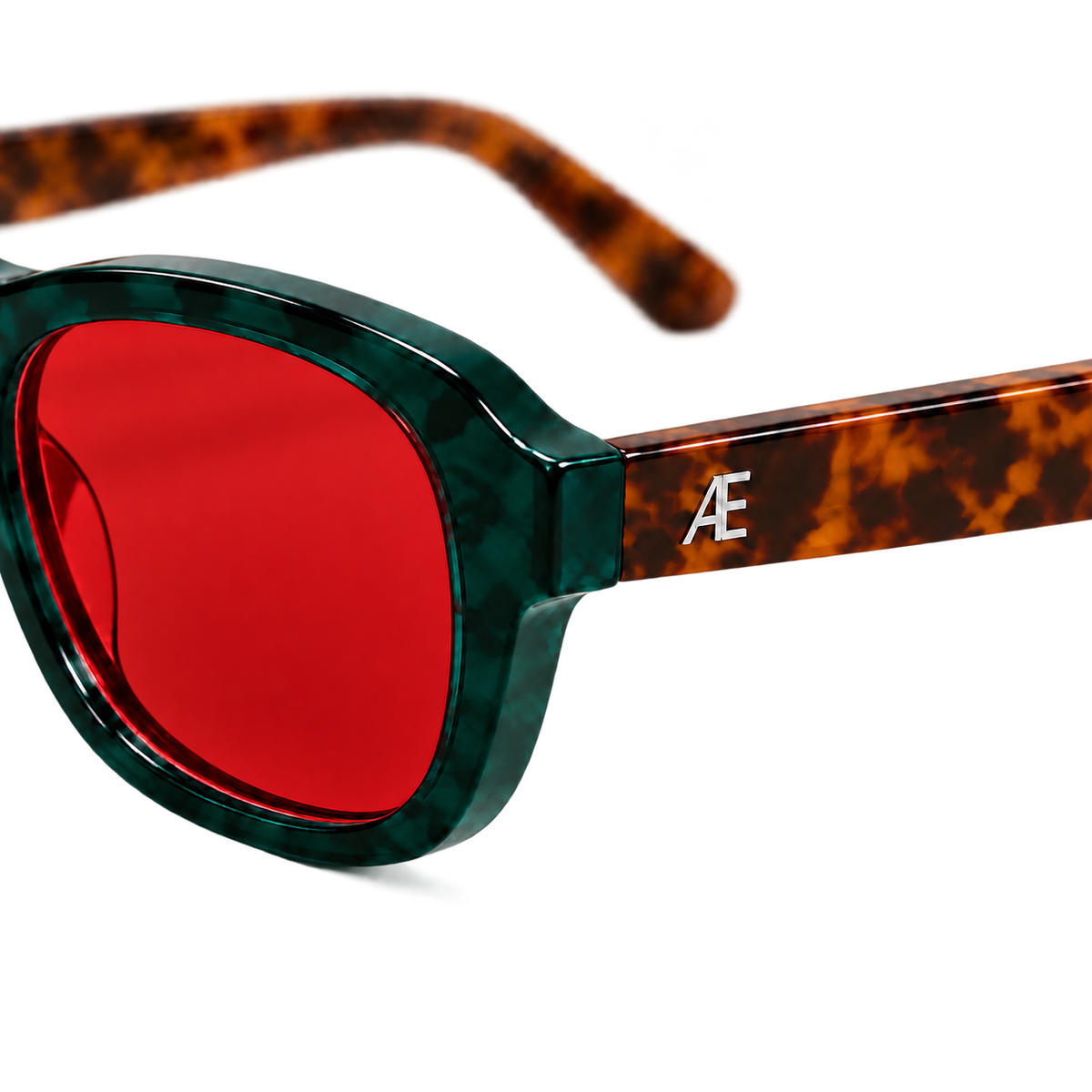 Macro view of the ATÉN Forest/Tortoise frame beside a deep red After Hours lens; deep forest-green marbled rim, brown tortoiseshell temple and silver ATÉN monogram, isolated with a soft natural shadow.
