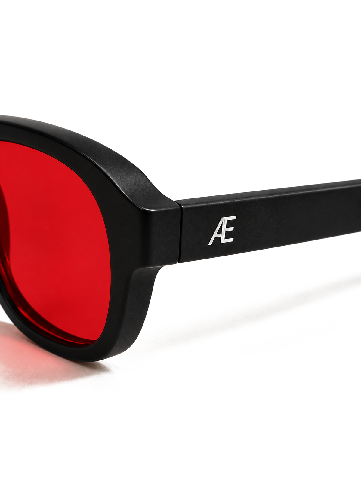 Transparent-background macro cutout of the ATÉN matte black outer hinge and temple beside the edge of a deep red After Hours lens; a silver ATÉN monogram is centred on the temple, with a soft red cast shadow beneath the frame.