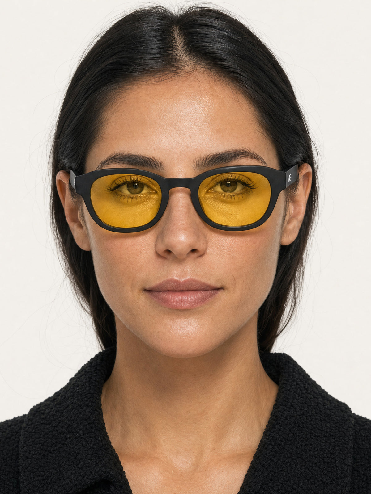 Straight-on head-and-shoulders studio portrait of a dark-haired woman looking at the camera while wearing ATÉN matte black glasses with pronounced amber Focus Mode lenses. Her straight hair is centre-parted and tucked behind her ears; she wears a black textured collared coat, and the silver ATÉN temple monogram is visible against a plain off-white background.