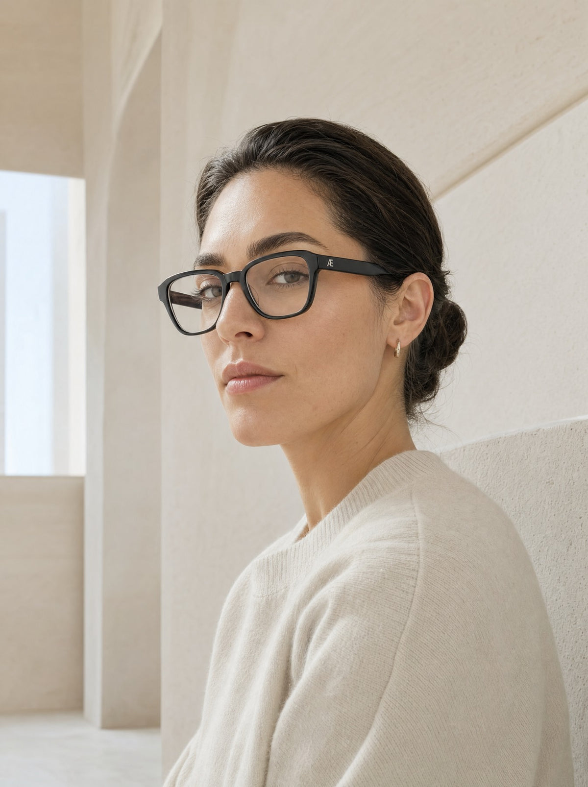 Three-quarter portrait of a dark-haired woman with her hair in a low bun, small gold hoop earrings and a cream knit sweater, wearing matte-black rectangular ATÉN frames with Everyday Mode lenses and a silver Æ temple monogram; she looks toward the camera against pale beige stone walls beside a bright window opening.