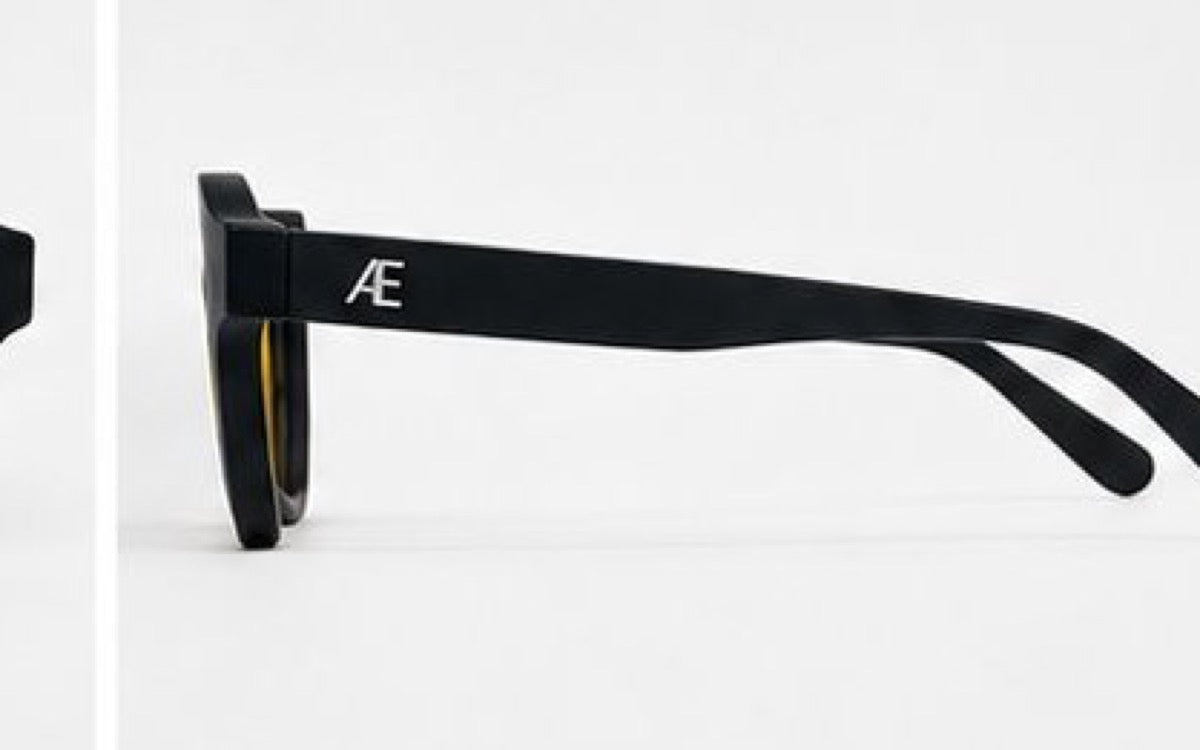 ATÉN matte black frame with a pronounced amber Focus Mode lens in a cropped left-side profile; black temple extends right with a silver ATÉN monogram near the hinge, on a white background.