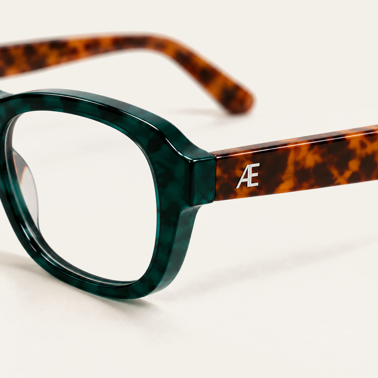 Close-up of the outer hinge and temple of ATÉN Forest/Tortoise glasses with an Everyday Mode lens; the deep forest-green marbled rim fills the left foreground, the brown tortoiseshell temple carries a silver ATÉN monogram, and the far temple is softly blurred on the cream background.