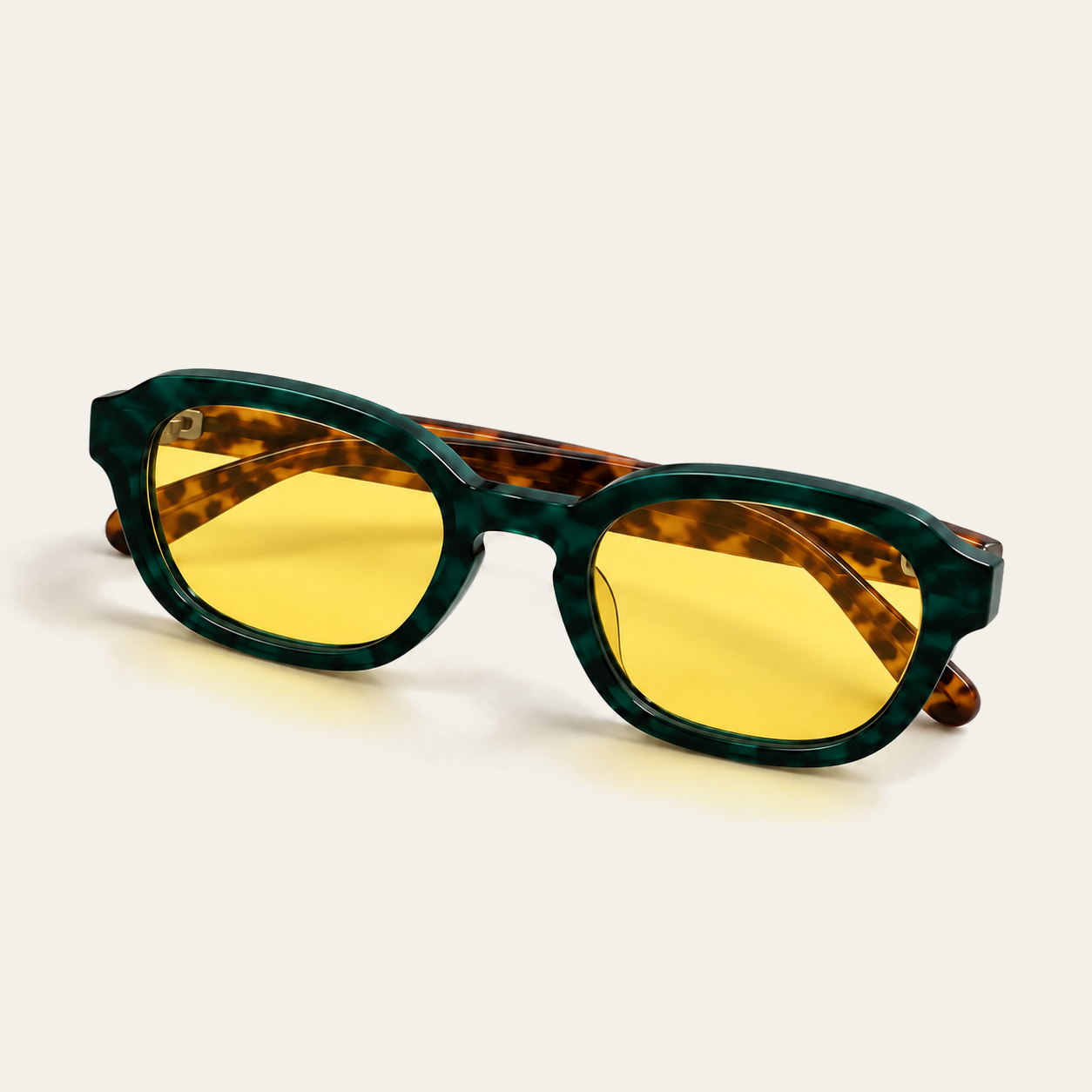 Folded ATÉN Forest/Tortoise glasses with pronounced amber Focus Mode lenses viewed from a raised front-left angle; the brown tortoiseshell temples overlap behind the deep forest-green marbled front, with amber reflections and a soft shadow on the cream background.