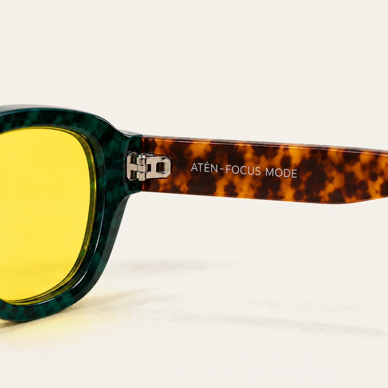 Close-up of the inner temple of ATÉN Forest/Tortoise glasses with a pronounced amber Focus Mode lens; the silver hinge is exposed, the brown tortoiseshell temple is engraved ATÉN · FOCUS MODE, and the frame casts a soft amber-tinted shadow on the cream background.