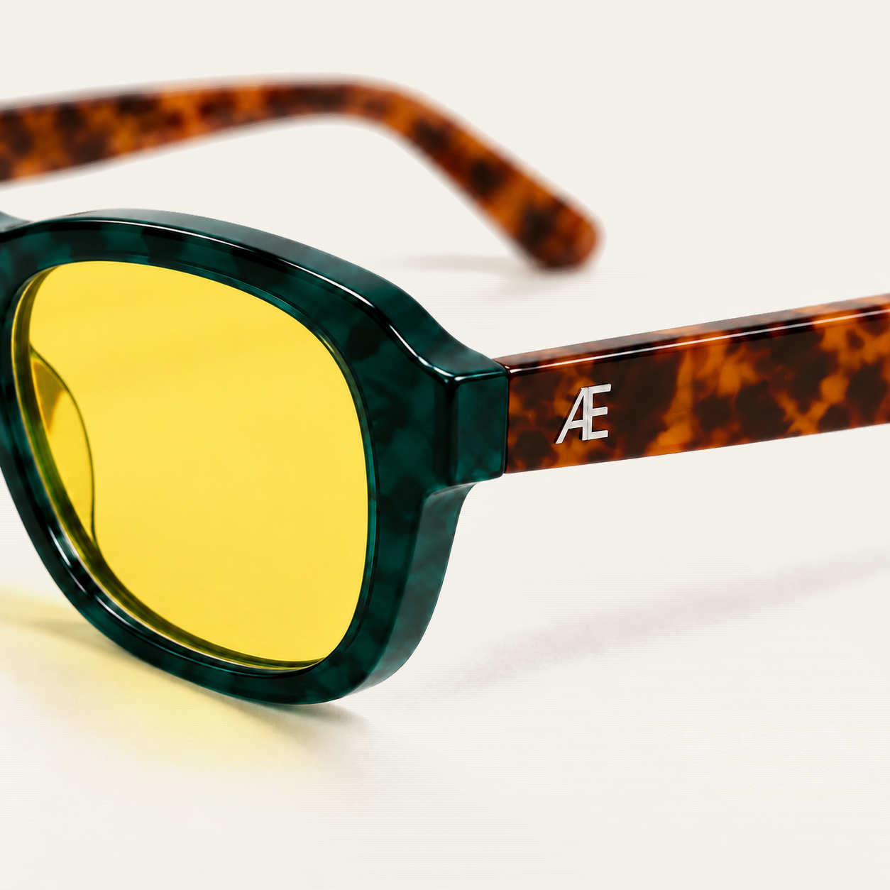 Close-up of the outer hinge and temple of ATÉN Forest/Tortoise glasses with a pronounced amber Focus Mode lens; the deep forest-green marbled rim and pronounced amber lens fill the left foreground, the brown tortoiseshell temple carries a silver ATÉN monogram, and the far temple is softly blurred on the cream background.