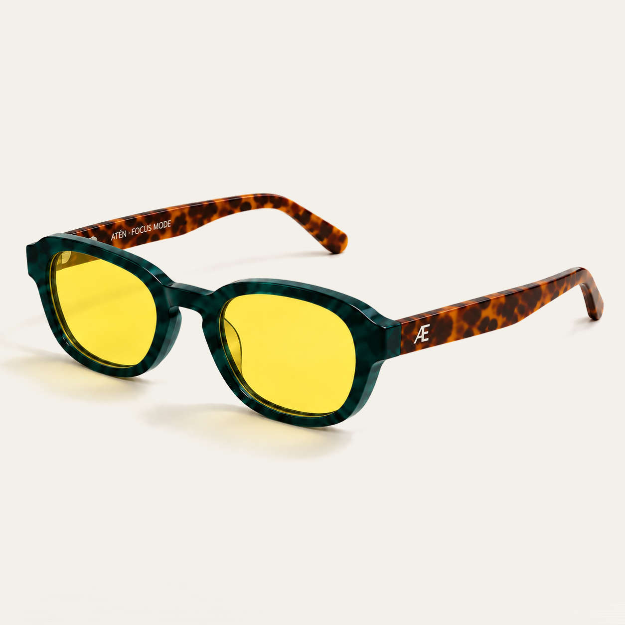 Open ATÉN Forest/Tortoise glasses with pronounced amber Focus Mode lenses in a front-right three-quarter view; the frame has a deep forest-green marbled front, brown tortoiseshell temples and a silver ATÉN monogram, with both temples open and amber-tinted shadows beneath the glasses on the cream background.